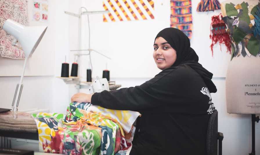 student smiling to camera sat at sewing machine with colourful cloth in textile room