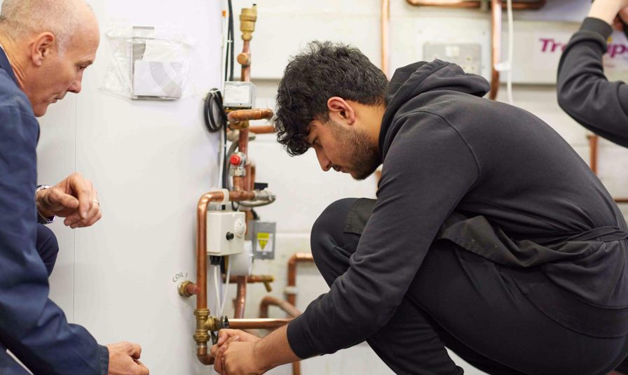 tutor watches over plumbing student as he turns a valve on pipes