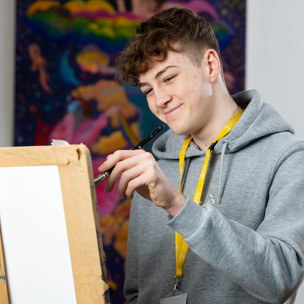 male student smiles whilst painting a canvas on an easel stood in front of a colourful painting