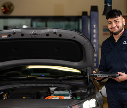 student in mechanics overalls smiling to camera holding diagnostic tool next to car with bonnet open