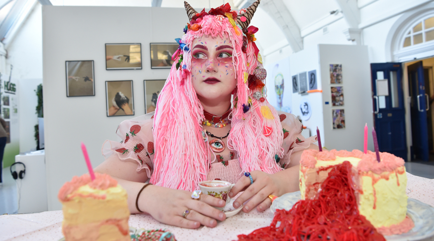 artist dressed with long pink hair and ice cream accessories with cakes on table holding a tea cup