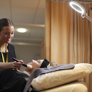 Student applying makeup onto another student lying down in a beauty room.