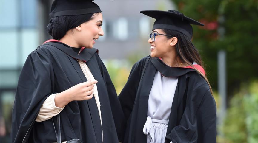 2 women in graduation cap and gowns smiling to each other