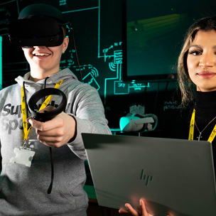 young man with VR headset and hand controllers stood next to young woman holding laptop