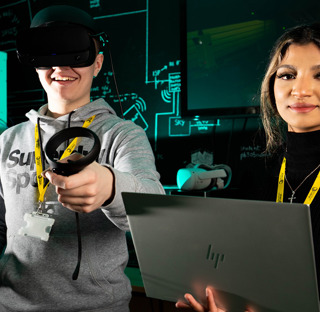 young man with VR headset and hand controllers stood next to young woman holding laptop