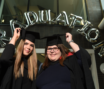 2 women in graduation cap and gowns cheering to camera under graduation balloons