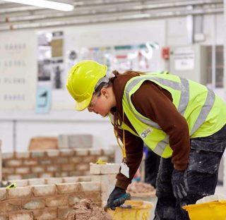 young woman in hard hat, hi-vis vest and safety goggles leaning over with trowel in mortar surrounded by brick walls