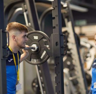 sports student stands with barbell weight whilst talking other student
