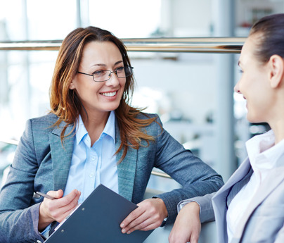 2 women talking on office settee smiling to each other 1 with clipboard and pen