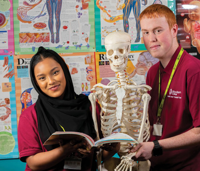 male and female student stood with open textbook and skeleton model in front of wall covered in anatomy posters