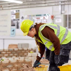 young man in hi-vis vest wearing hard hat places mortar on brick wall in workshop