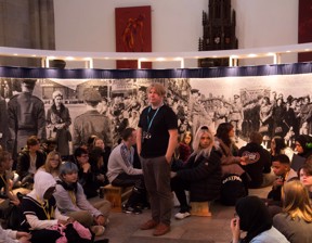 group of students sat around tutor Jamie Holman in front of large artwork of the queen