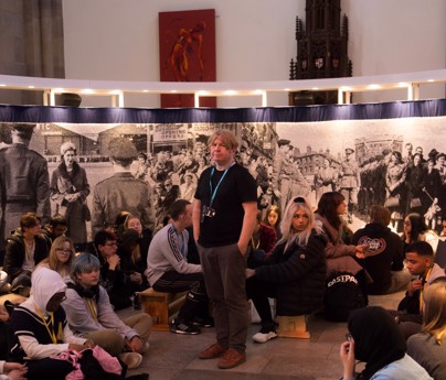 group of students sat around tutor Jamie Holman in front of large artwork of the queen