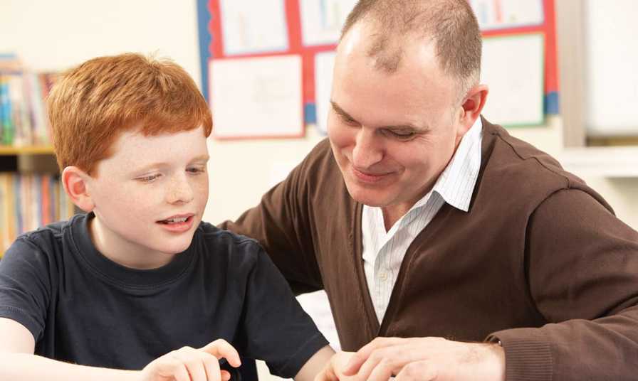 male teaching assistant sits with school child looking at book