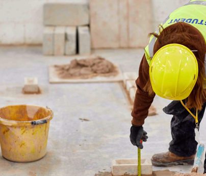 young man in hi-vis vest using trowel to level brick on wall in workshop
