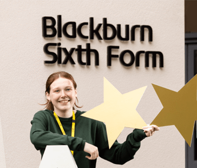 Ellie Driver smiles holding 2 gold stars in front of Blackburn Sixth Form sign