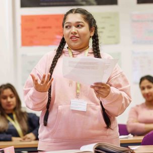 Student stood up in classroom reading from a piece of paper wearing a pink hoodie.