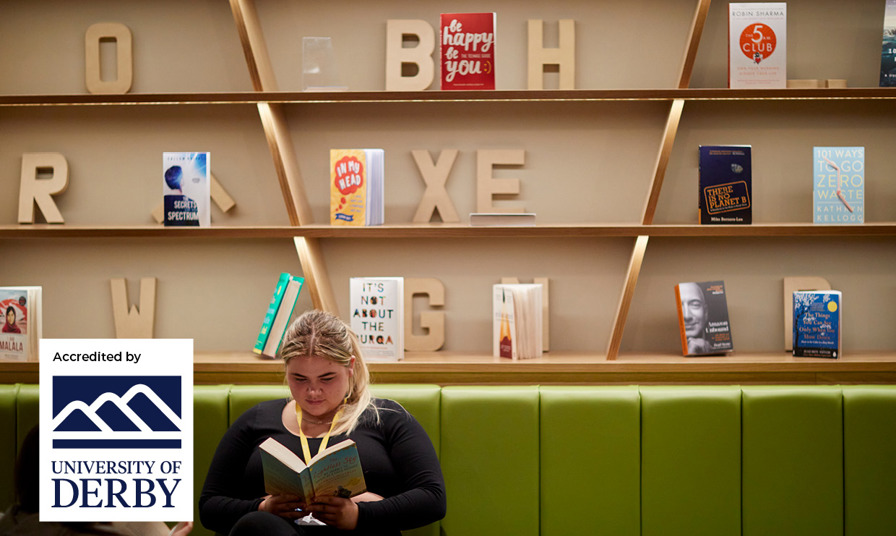 students sat in the Exchange learning zone reading a book. A bookshelf is behind her with University of Derby logo