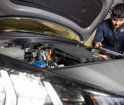 student in mechanics overalls looking at diagnostic tool over open bonnet of car