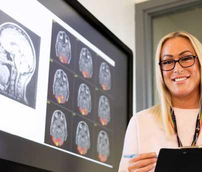 female student smiles to camera holding clip board stood next to screen with images of brain scans