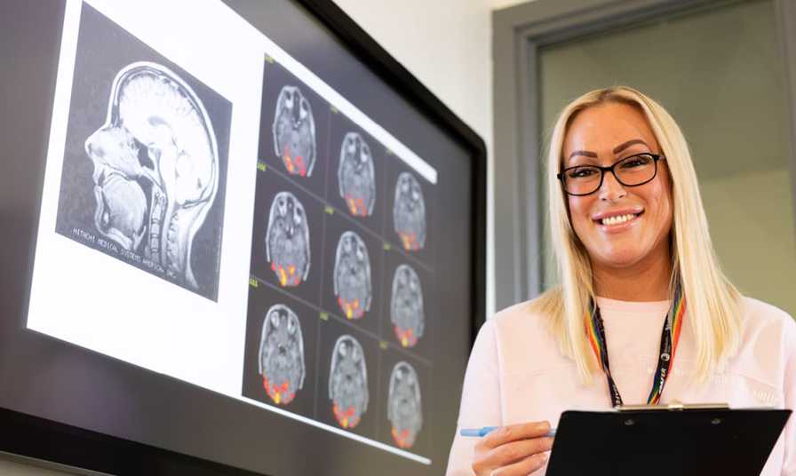 female student smiles to camera holding clip board stood next to screen with images of brain scans