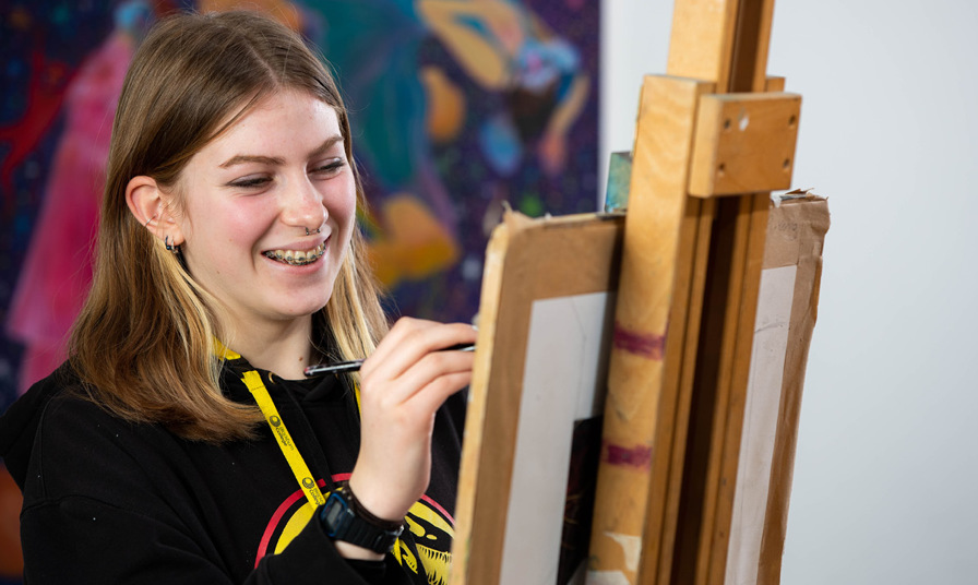 student smiling whilst drawing on easel stood in front of colourful painting