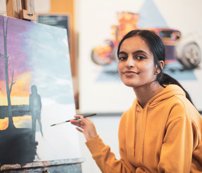 student smiles to camera whilst painting colourful picture on easel