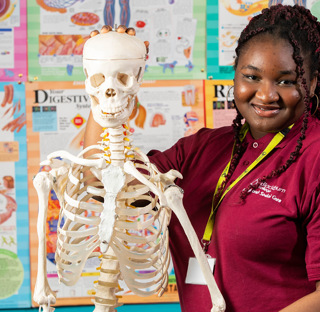 female health student smiling to camera holding model skeleton in front of wall covered with anatomy posters