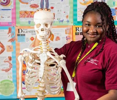 female health student smiling to camera holding model skeleton in front of wall covered with anatomy posters