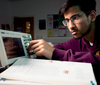 male student turning dials on oscilloscope with open text book