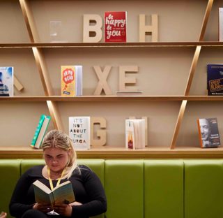 student sits reading book on long green sofa in front of book casing with scattered books and 3d wooden lettering