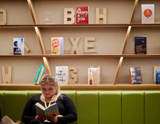 student sits reading book on long green sofa in front of book casing with scattered books and 3d wooden lettering