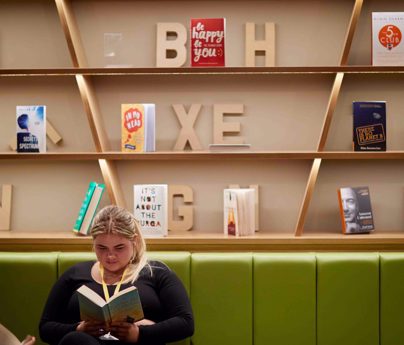 student sits reading book on long green sofa in front of book casing with scattered books and 3d wooden lettering