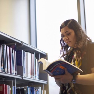 student looks through book stood next to library shelf