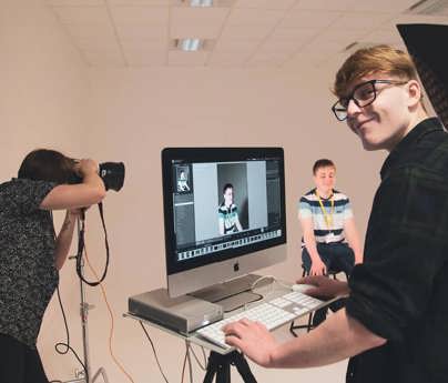 student smiles to camera working on mac whilst photo is taken of model in studio