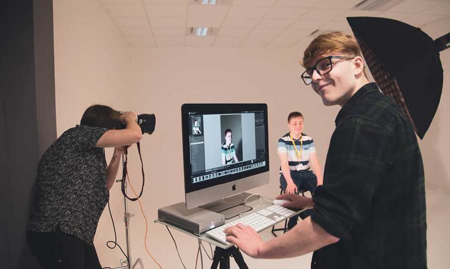 student smiles to camera working on mac whilst photo is taken of model in studio