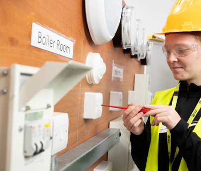 female electrical student using screwdriver on light switch casing on wall with various electrical fittings 