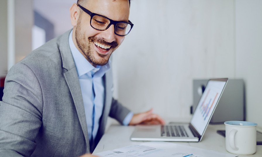 smiling man leans over to read figures on paper whilst working on laptop in office