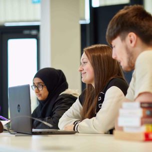 Sixth form student sitting with classmates smiling with a laptop on the desk.