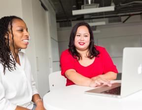 2 woman sat at desk whilst one shows something on a laptop