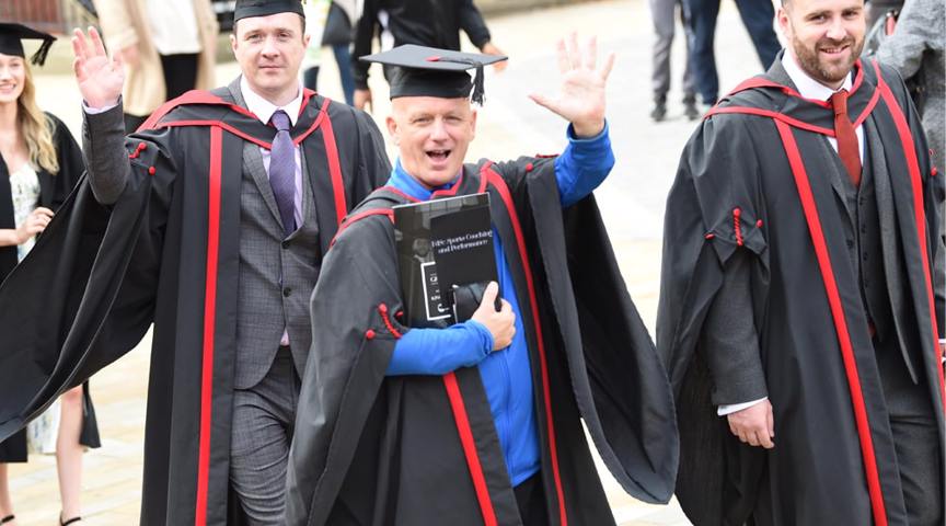 3 men in graduation cap and gowns smiling and waving to camera