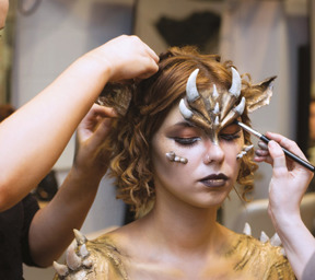 2 women applying makeup and facial accessories and gold paint to model