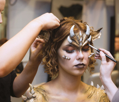 2 women applying makeup and facial accessories and gold paint to model